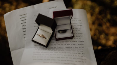 two boxes with wedding bands, one gold and the other brown, sitting onto of wedding vow papers