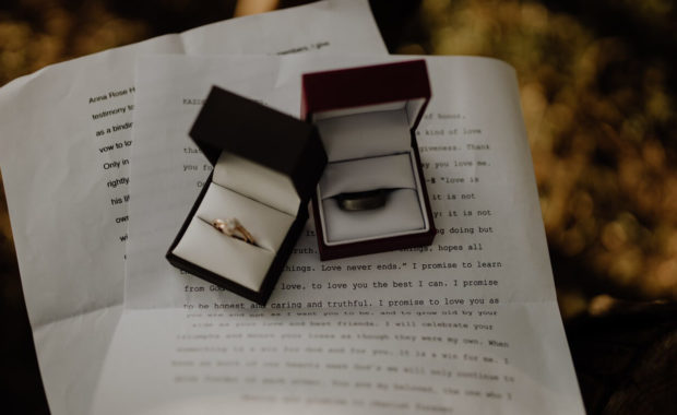 two boxes with wedding bands, one gold and the other brown, sitting onto of wedding vow papers