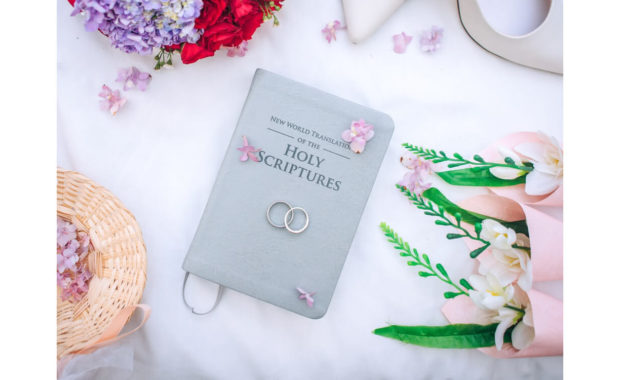 picture of two gold rings sitting on top of a holy scriptures book with blowers surrounding it on a white cloth