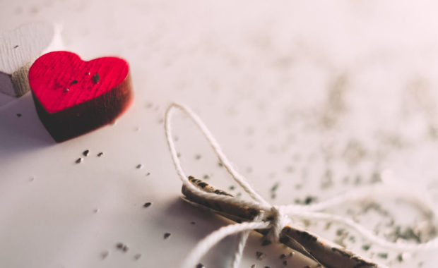 red and white heart laying on white table with sparkles around them and a stick with a box tied on it